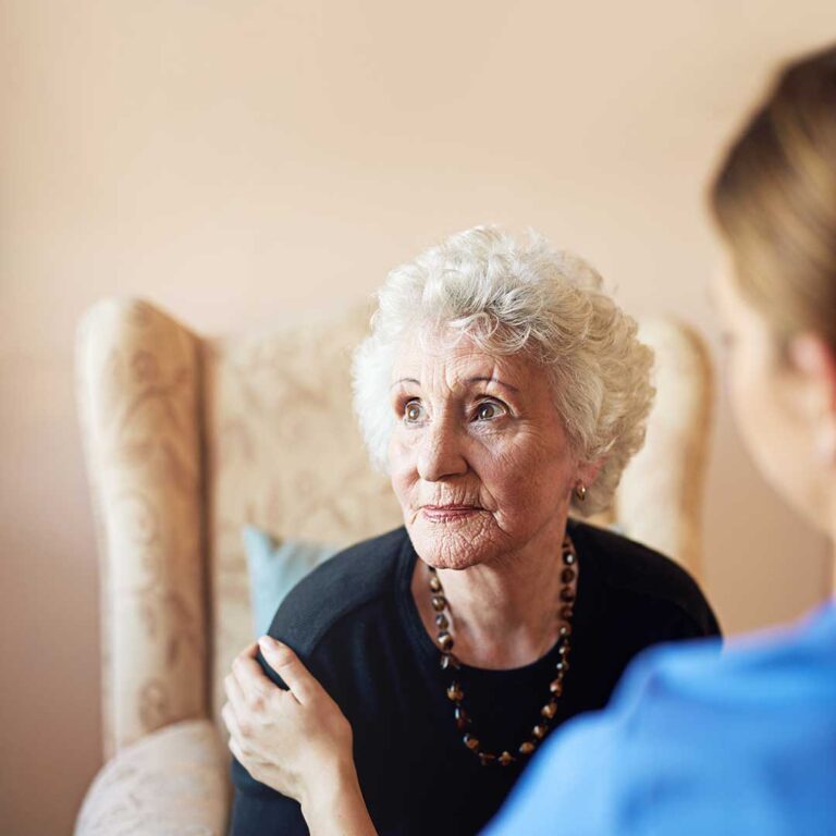 Two elderly residents sharing a meal and talking with staff at a communal dining table, with personal items visible in the background.