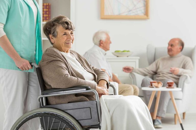 Older adults playing cards and chatting at a shared table in a sunny, home-like common area.